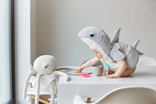 A toddler sits in a shark costume on a table and gives a stuffed animal a flip.